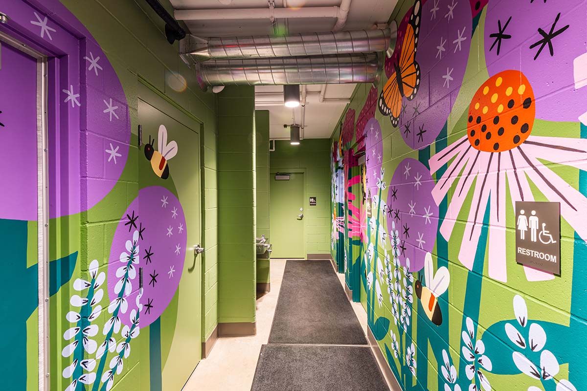 Colorful mural hallway with green walls, flowers, and a butterfly, leading to a door at the end and a restroom sign on the right wall.