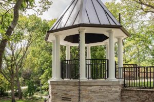 Octagonal white gazebo with columns and black railings on a stone pedestal in a lush, tree-filled park.