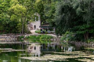 Stone building with arched doorway and a gazebo tucked among trees beside a calm pond with floating lily pads.