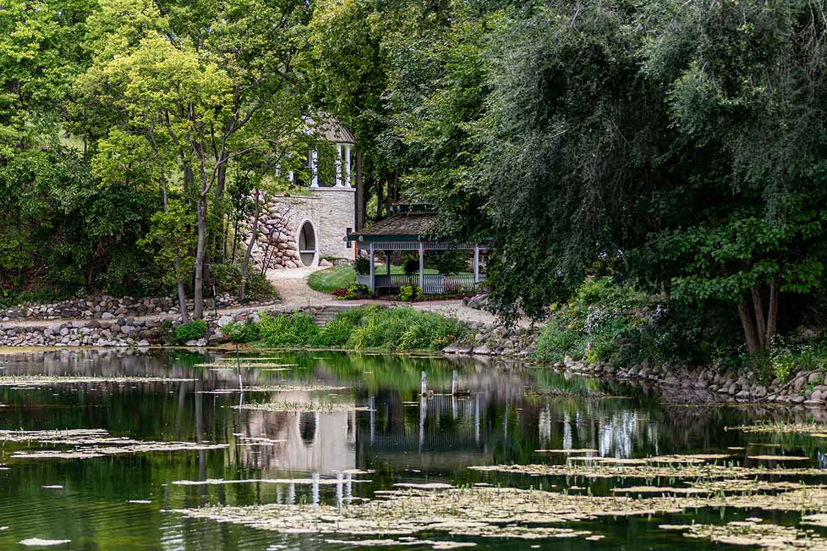 Stone building with arched doorway and a gazebo tucked among trees beside a calm pond with floating lily pads.