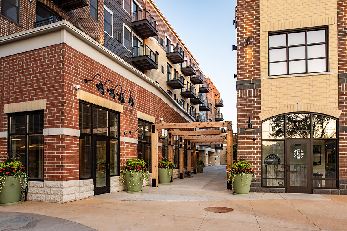 Corner view of Middleton Center brick building with pergola entry, upper-floor balconies, and retail storefronts at street level