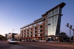 Street view of Peloton Residences at dusk with brick facade, glass curtain wall tower, and red awnings at street level