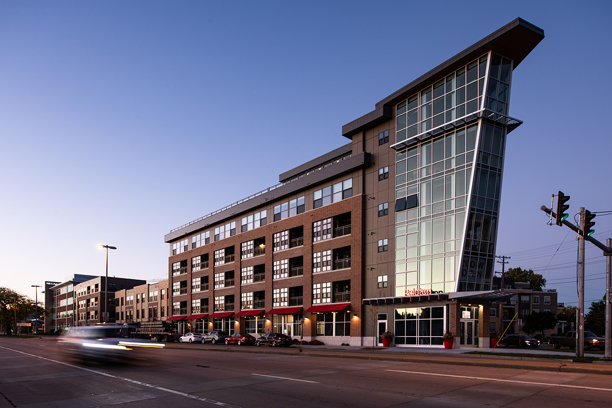 Street view of Peloton Residences at dusk with brick facade, glass curtain wall tower, and red awnings at street level