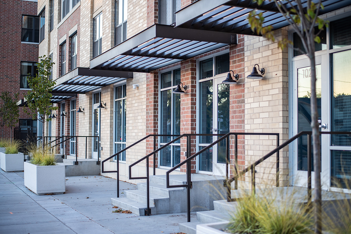 Street-level entry at Peloton Residences with metal canopy, brick and tan facade, and concrete planters