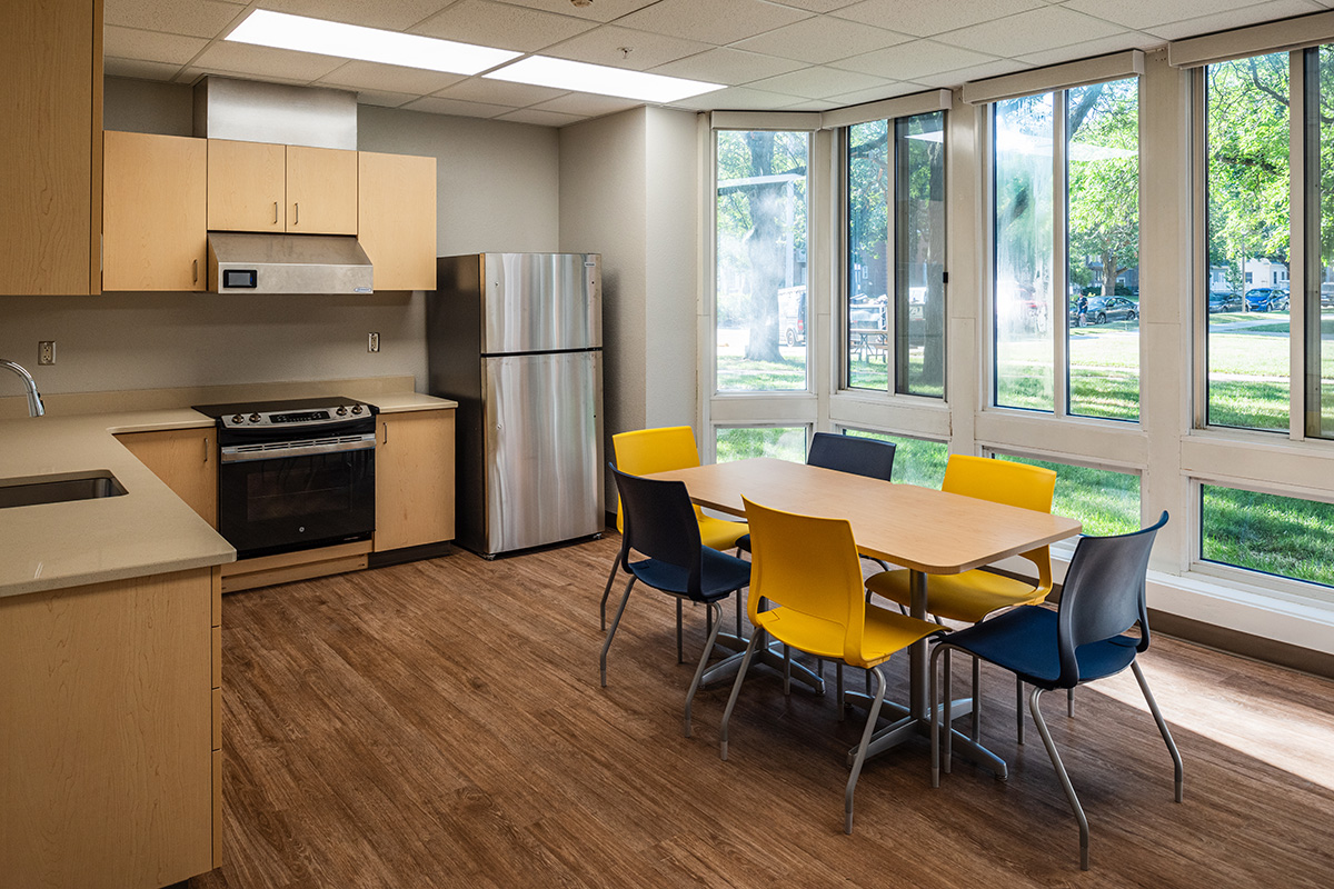Residential kitchen and dining area at Beloit College Whitney Hall with wood cabinetry, yellow chairs, and large windows overlooking campus