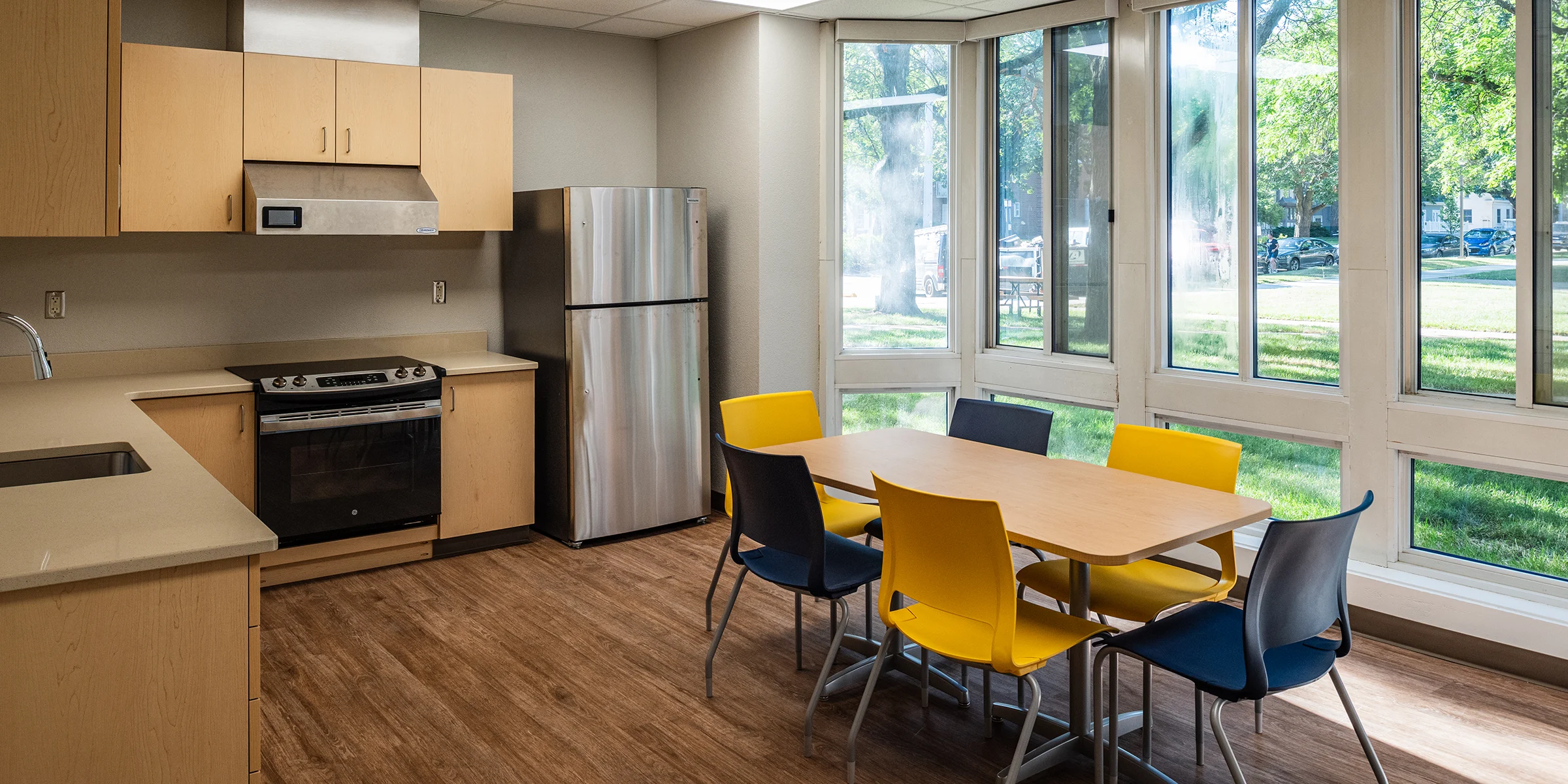 Residential kitchen and dining area at Beloit College Whitney Hall with stainless steel appliances, wood cabinetry, yellow and navy chairs, and large windows overlooking campus lawn