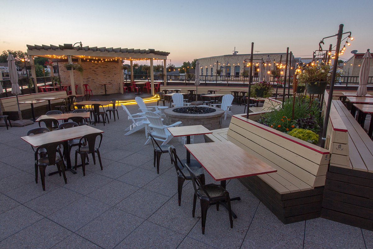 Wide view of rooftop patio at Village Crossing at dusk with dining tables, fire pit, pergola, and string lights