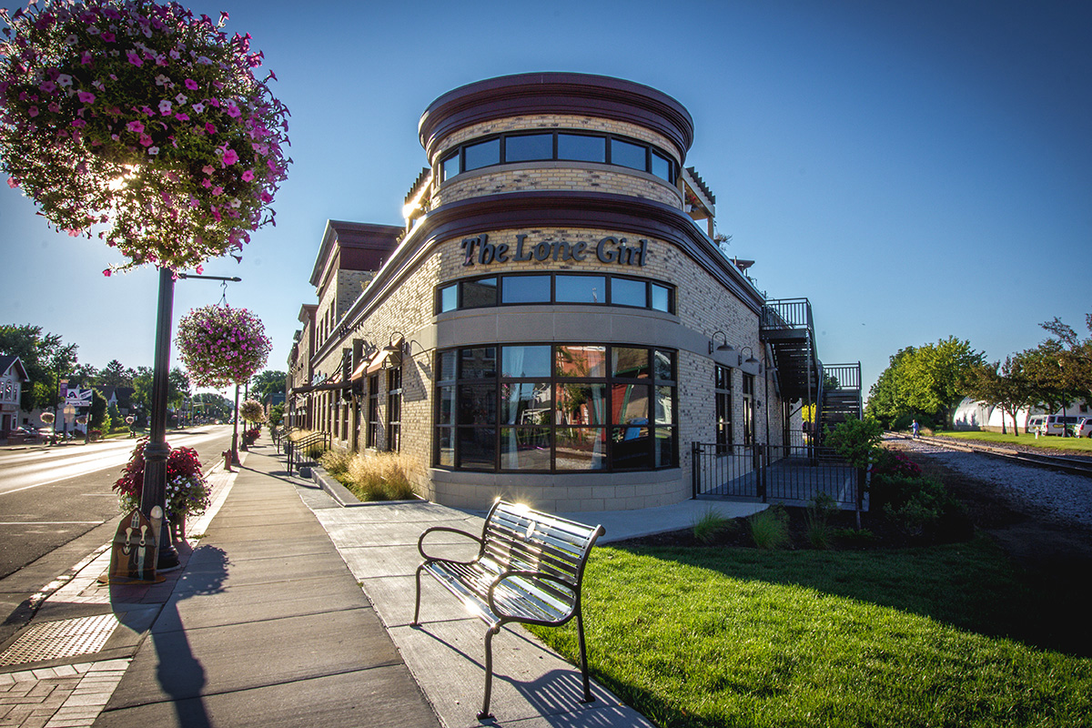 Street-level corner exterior of The Lone Girl at Village Crossing with curved brick and tan facade, rooftop patio, and sidewalk landscaping